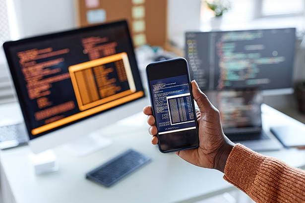 Personne tenant un téléphone portable devant un écran d’ordinateur alors qu’elle travaille au développement d’applications personnalisées pour une entreprise