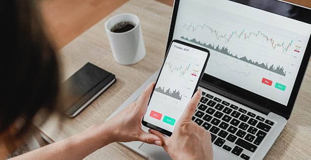 Une femme, spécialisée dans le développement d’applications mobiles, utilise un smartphone pour consulter un graphique boursier sur un ordinateur portable